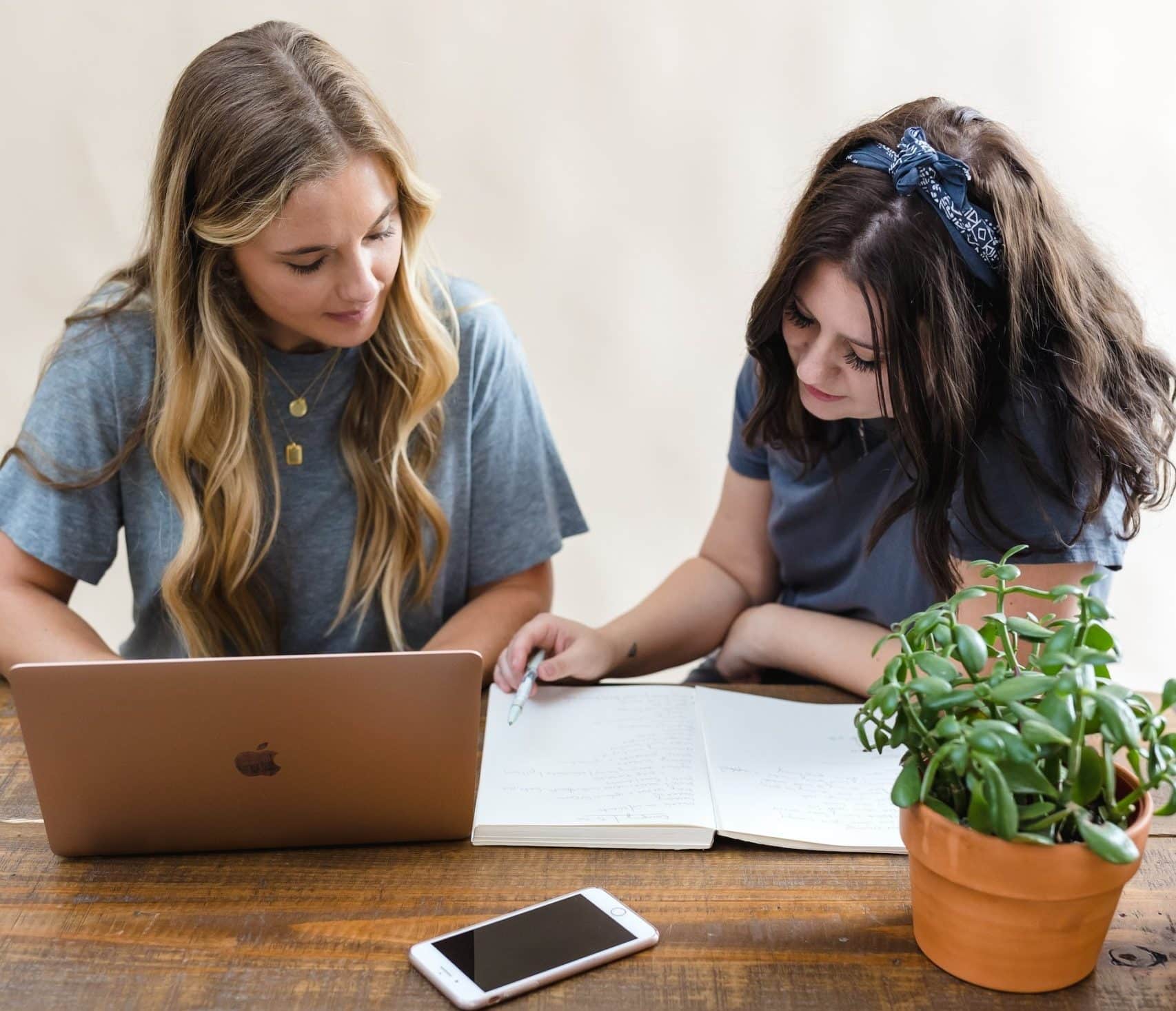 Two girls studying English at a wooden table, looking at an open book and laptop. One smartphone is put on the table.